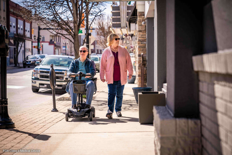 Two people walking outdoors, one using a mobility scooter, showcasing mobility products in action.