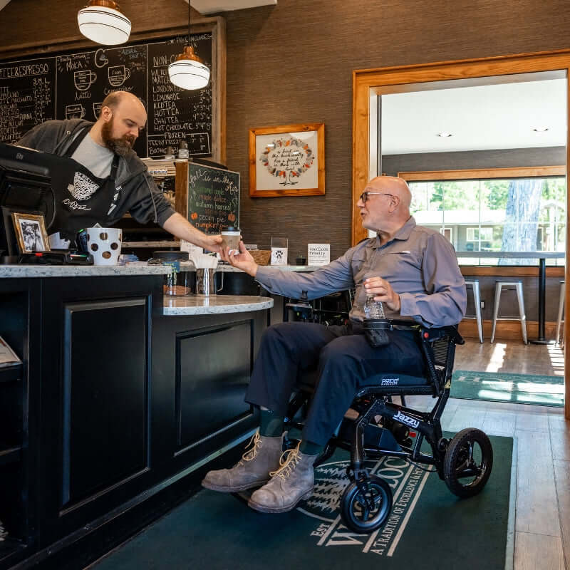 Senior man in Jazzy Ultra Light wheelchair enjoying coffee at cafe, showcasing the chair's design and portability.